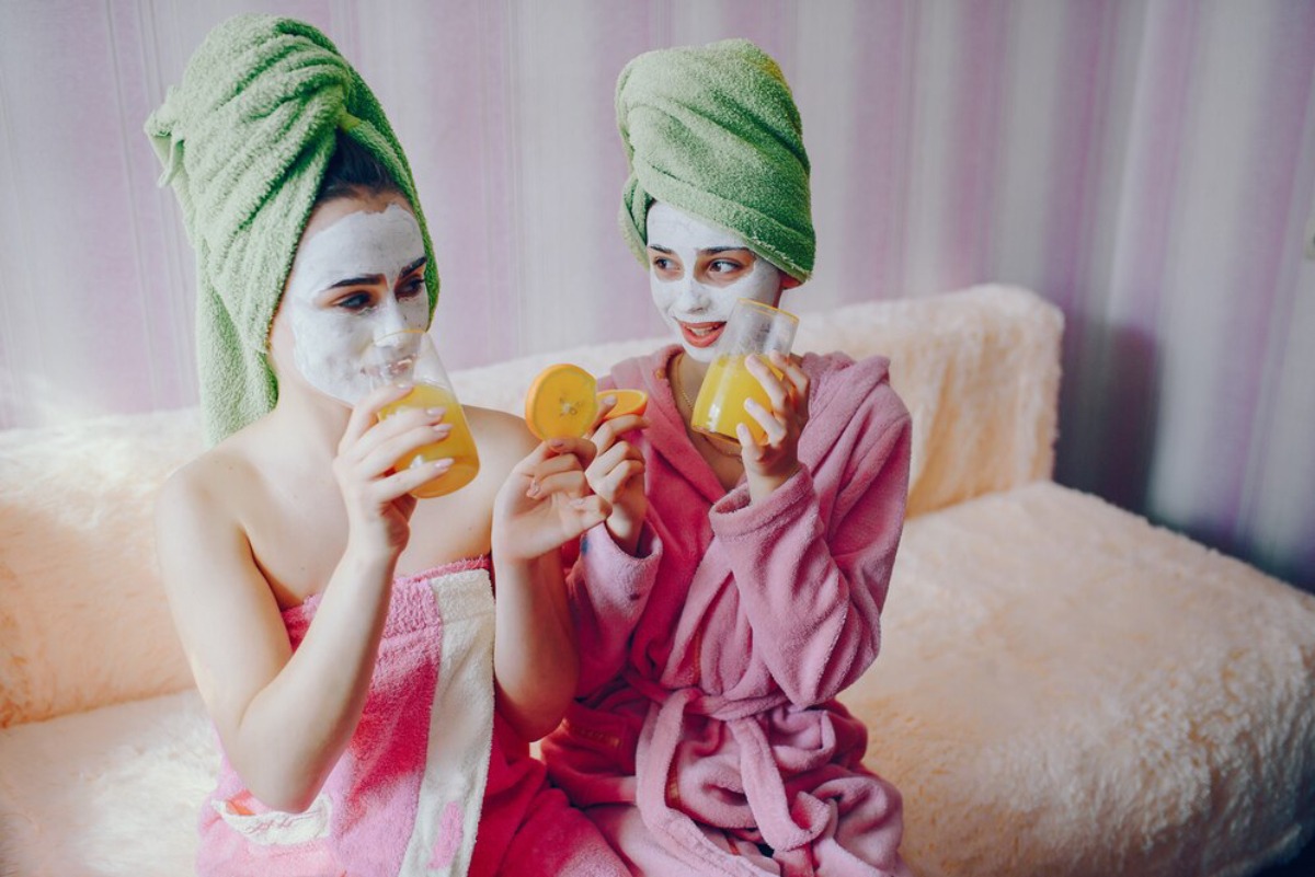 Two women in towels and robes pose cheerfully while holding refreshing drinks, sitting on a plush couch in a pastel setting.