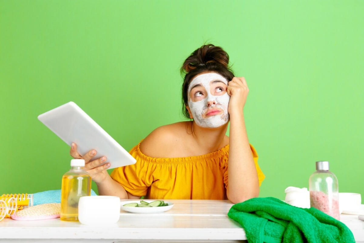 A person in an off-shoulder yellow top sits at a table with a tablet, cucumbers, towels, and various cosmetic bottles against a green background.