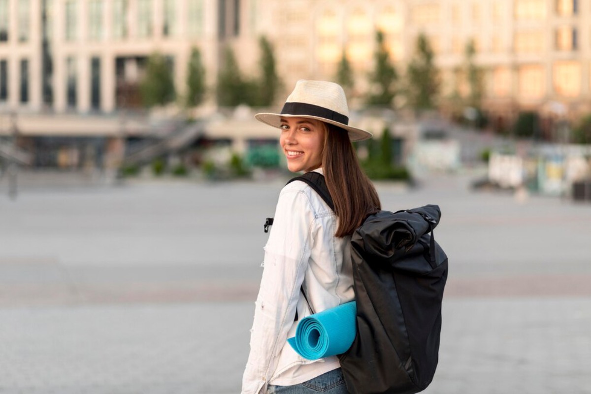 A woman wearing a straw hat and a white shirt carries a black backpack and a blue yoga mat in an urban setting.