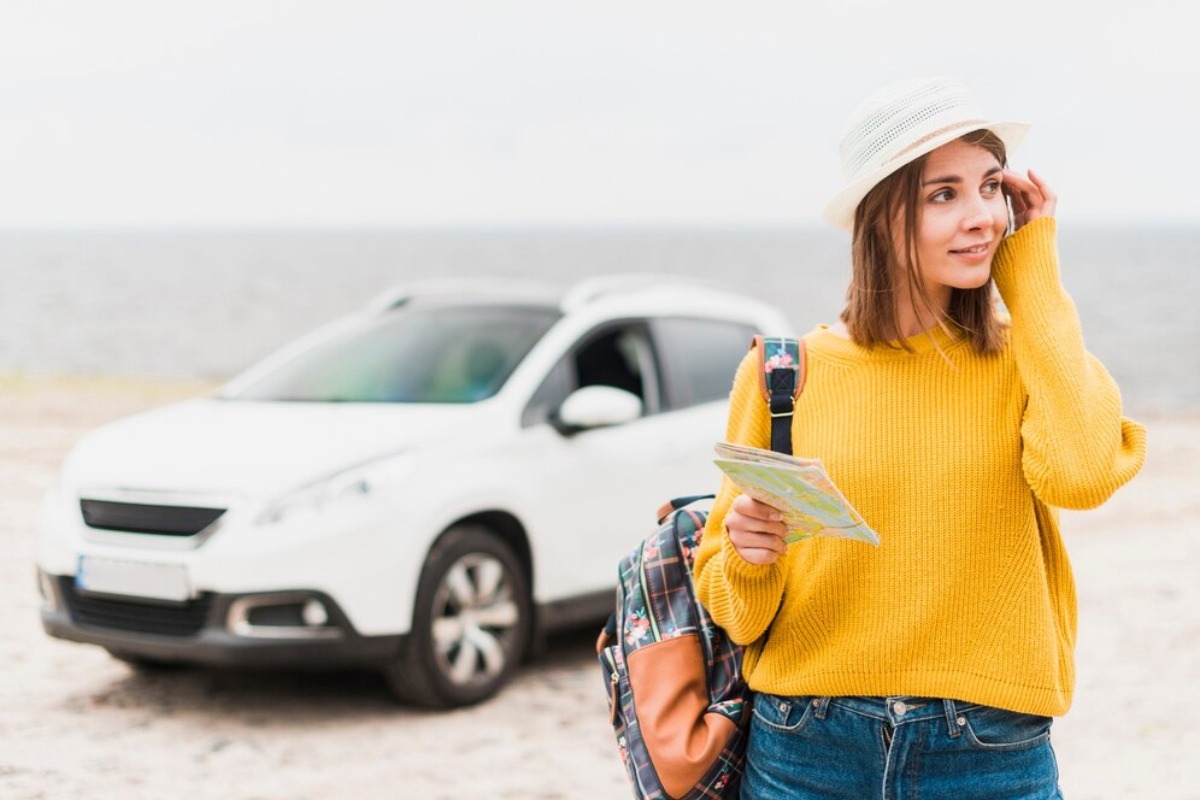 A person in a yellow sweater stands by a white car, holding a map, with a beach and ocean in the background.