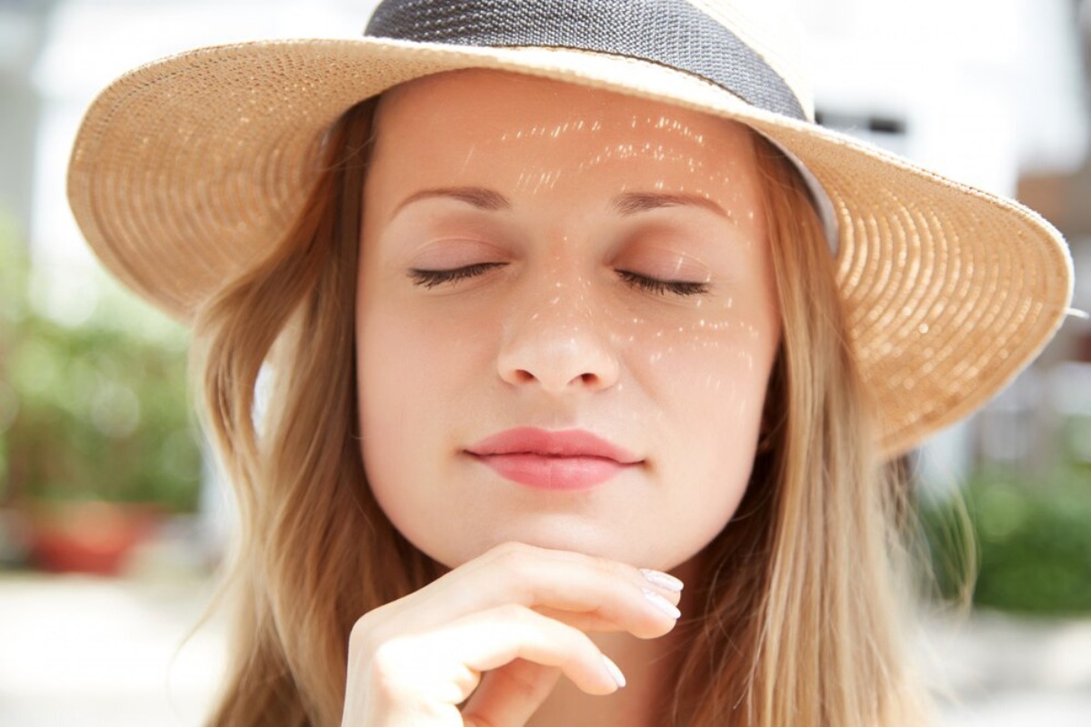 A woman with long hair wears a wide-brimmed hat, holding her hand near her mouth, with greenery blurred in the background.