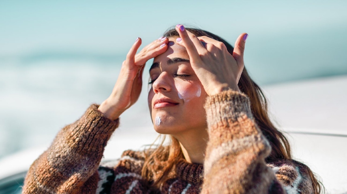 A person with long hair, wearing a cozy knit sweater, holds their hands to their hair against a backdrop of a blue sky and ocean.