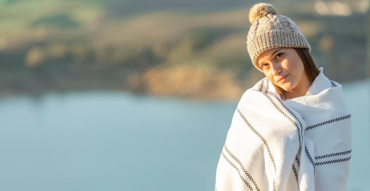 A person wrapped in a cozy blanket and wearing a knitted hat stands by a serene body of water, surrounded by lush hills.
