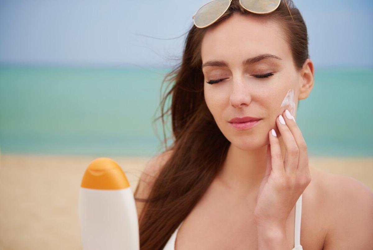 A woman in a white bikini holding a sunscreen bottle, with a beach and turquoise sea blurred in the background.