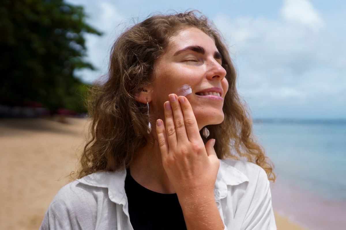 A person with curly hair stands on a beach, wearing a black top and a white shirt, gazing towards the ocean under a cloudy sky.