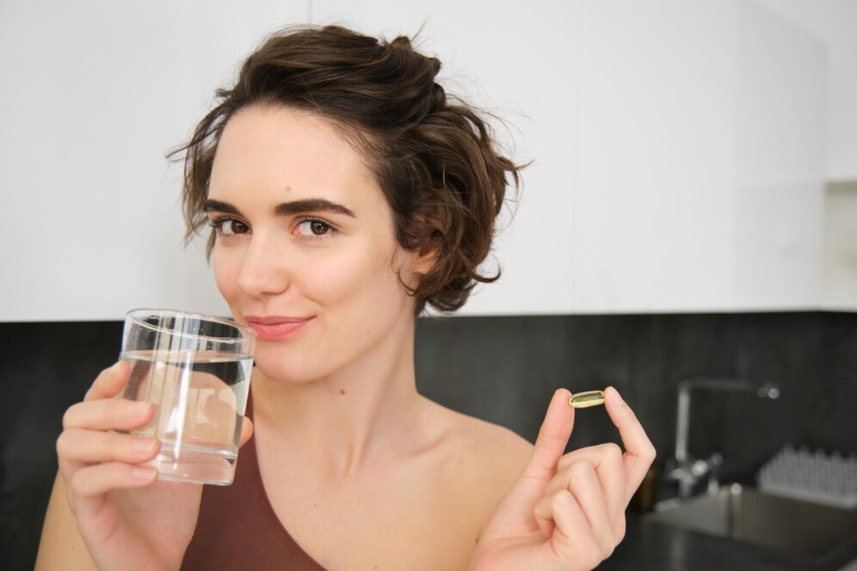 A person holding a glass of water in one hand and a capsule in the other, seated in a modern kitchen setting.