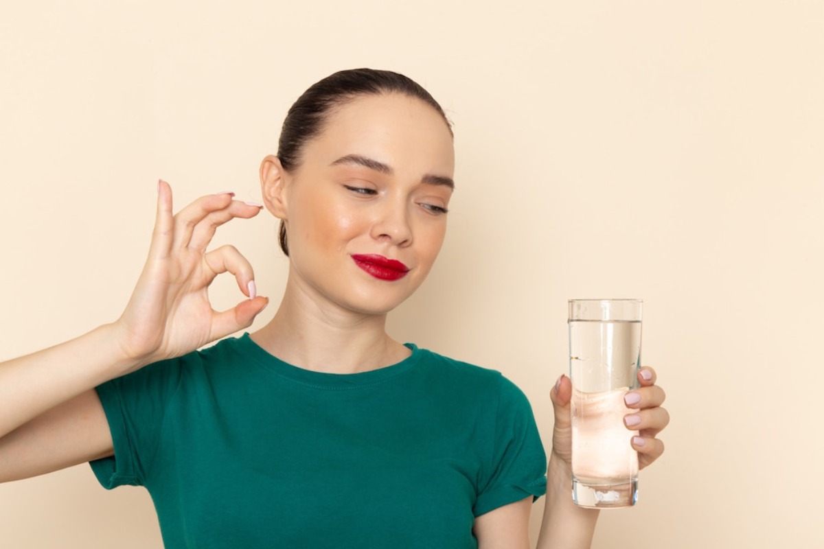 A person in a green shirt holds a clear glass of water in one hand and makes an OK gesture with the other against a beige background.