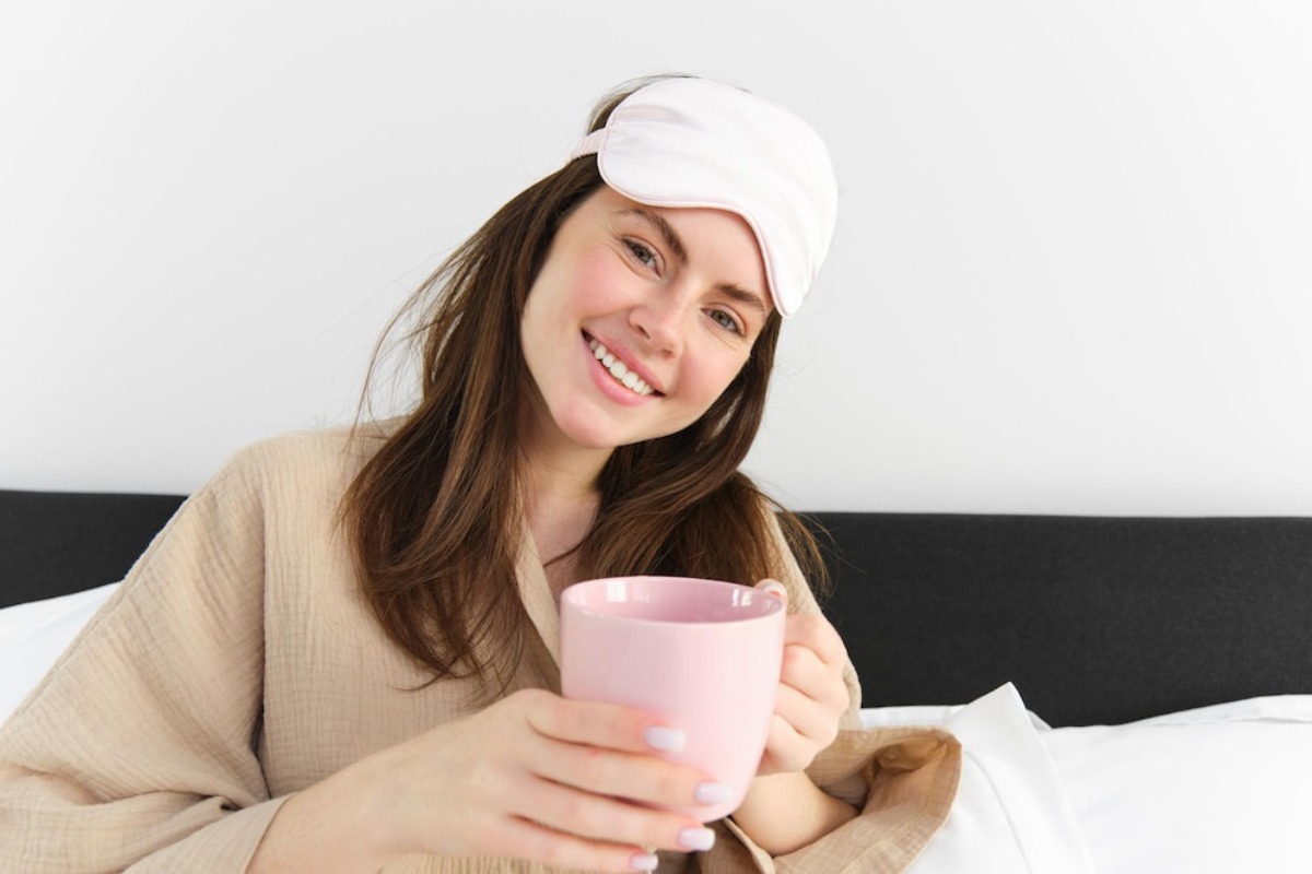 A woman in a cozy beige robe holds a pink mug, sitting comfortably on a bed with white linens against a minimalist background.