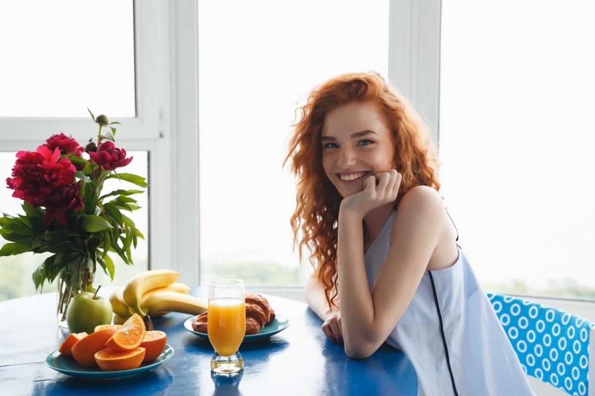 A woman sits at a blue table with a variety of fruits, a vase of red flowers, and a glass of orange juice. Bright, airy background.