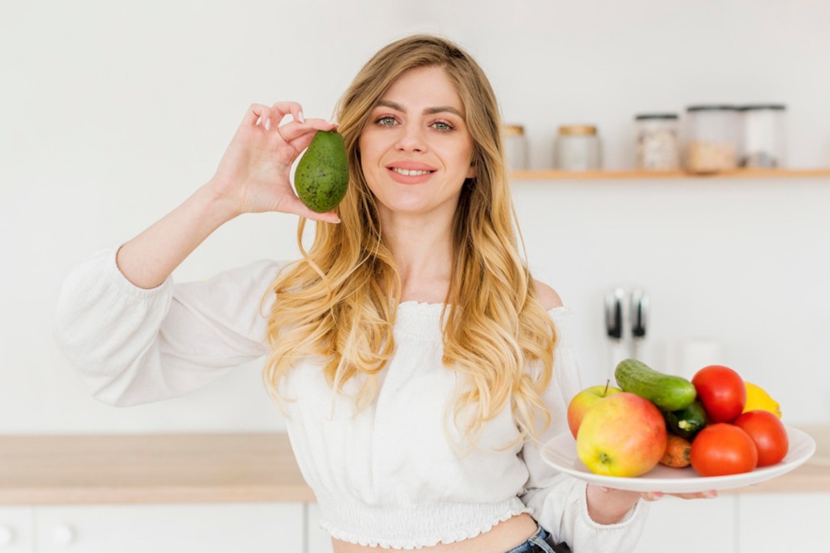A person holds an avocado in one hand and a plate of colorful fruits and vegetables in the other, standing in a bright kitchen.
