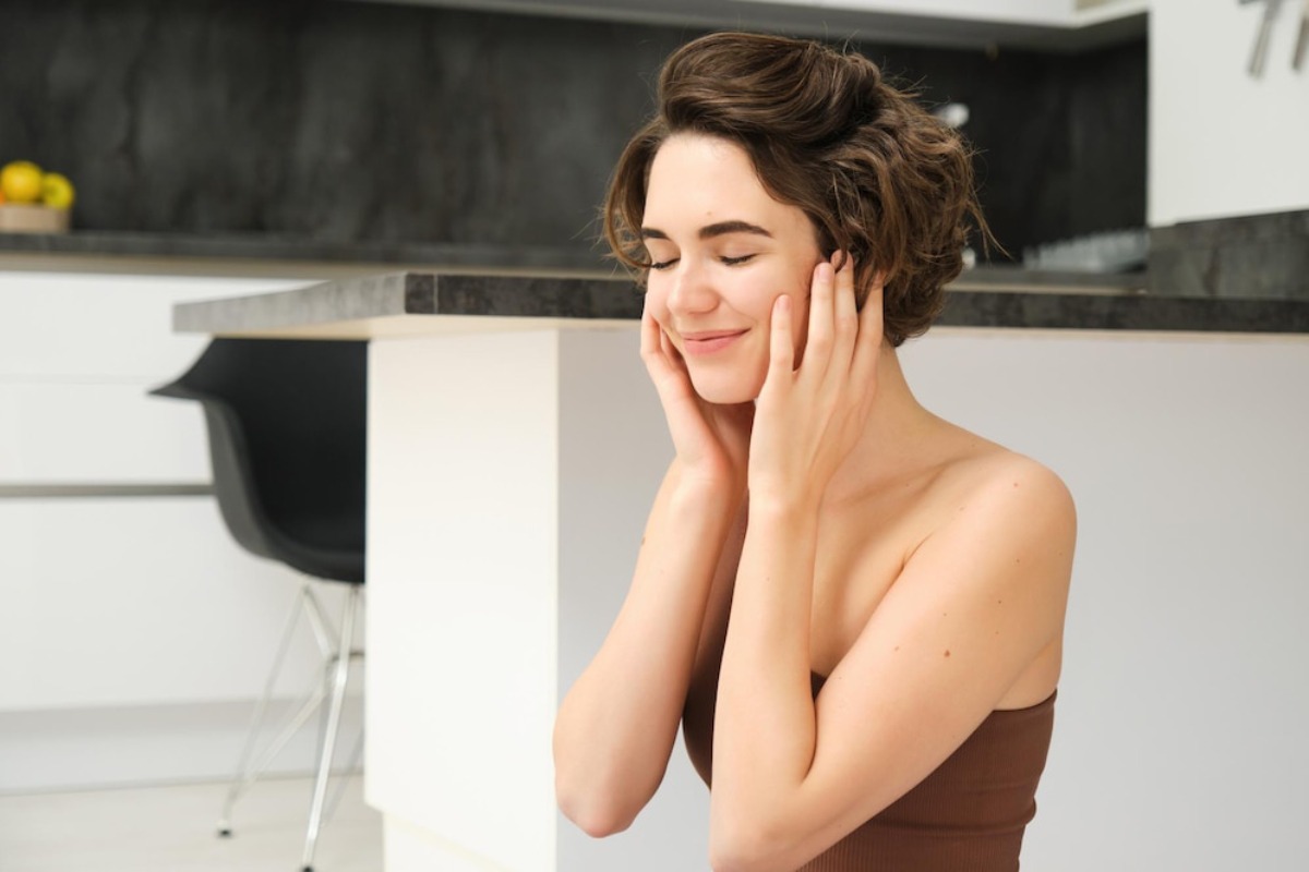 A woman with short, wavy hair sits on the floor, holding her head with both hands, in a modern kitchen setting.