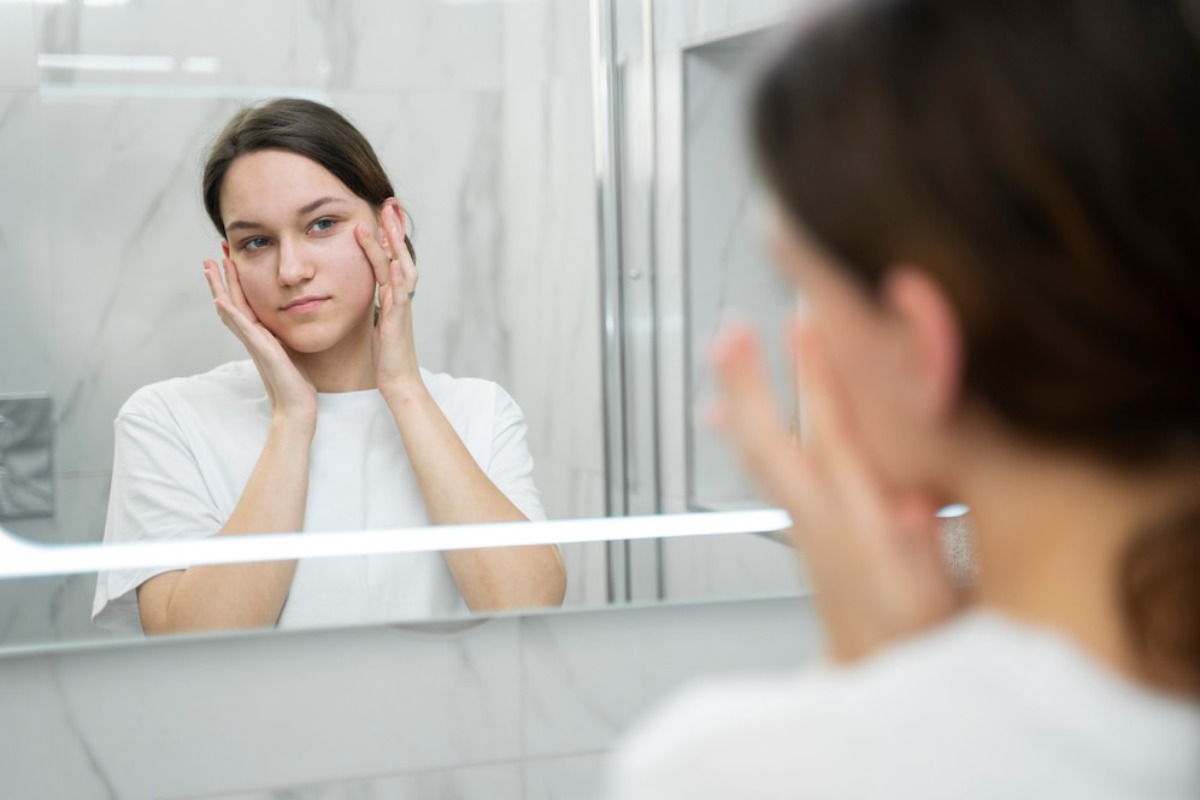 A person wearing a white shirt is applying skincare products while looking in a bathroom mirror.