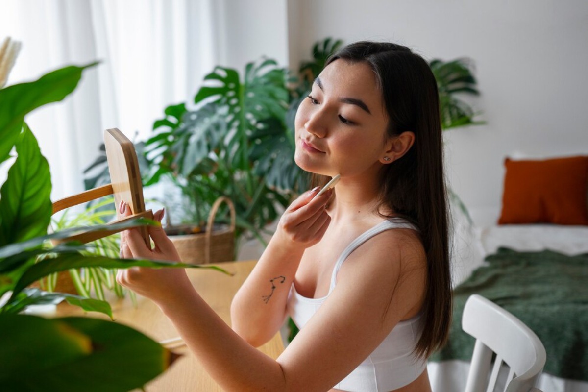 A woman in a white tank top applies makeup while sitting at a table surrounded by greenery, with a cozy bedroom in the background.