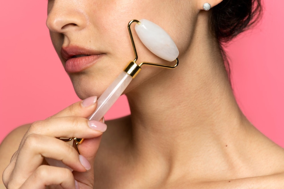 A person demonstrating facial skincare with a jade roller against a pink background, highlighting health and beauty practices.