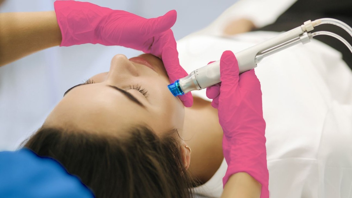 A beautician with pink gloves performs a facial treatment on a relaxed woman, using a microneedling device.