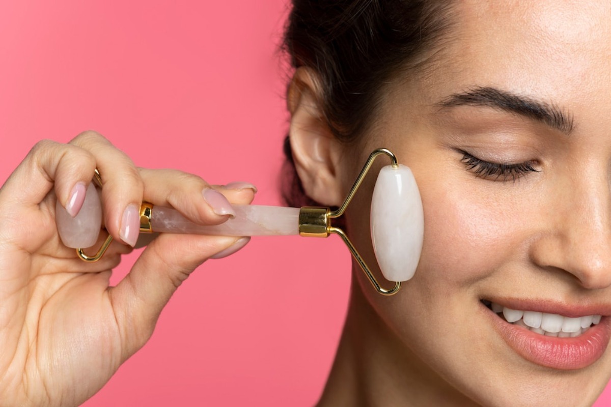 A hand holding a rose quartz facial roller against a pink background, demonstrating a skincare routine.