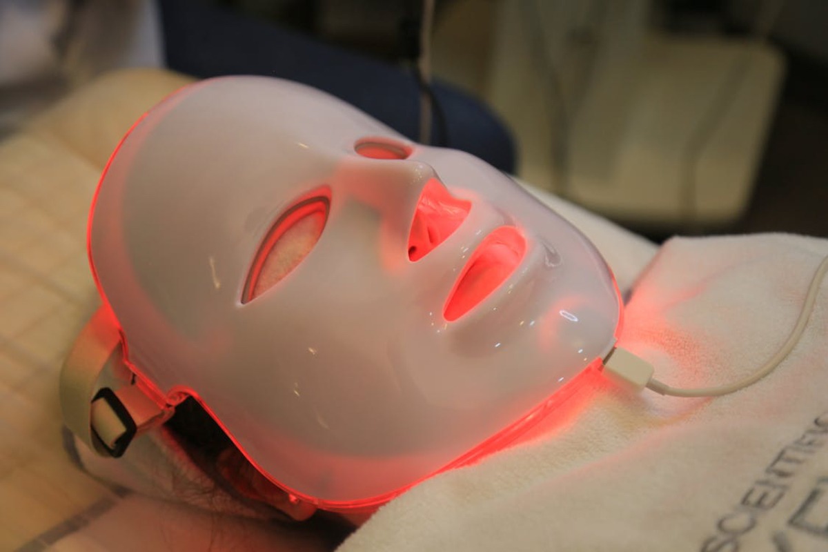 A red LED light therapy device positioned on a towel, with a white cable plugged into it, suggesting a skincare treatment setup.