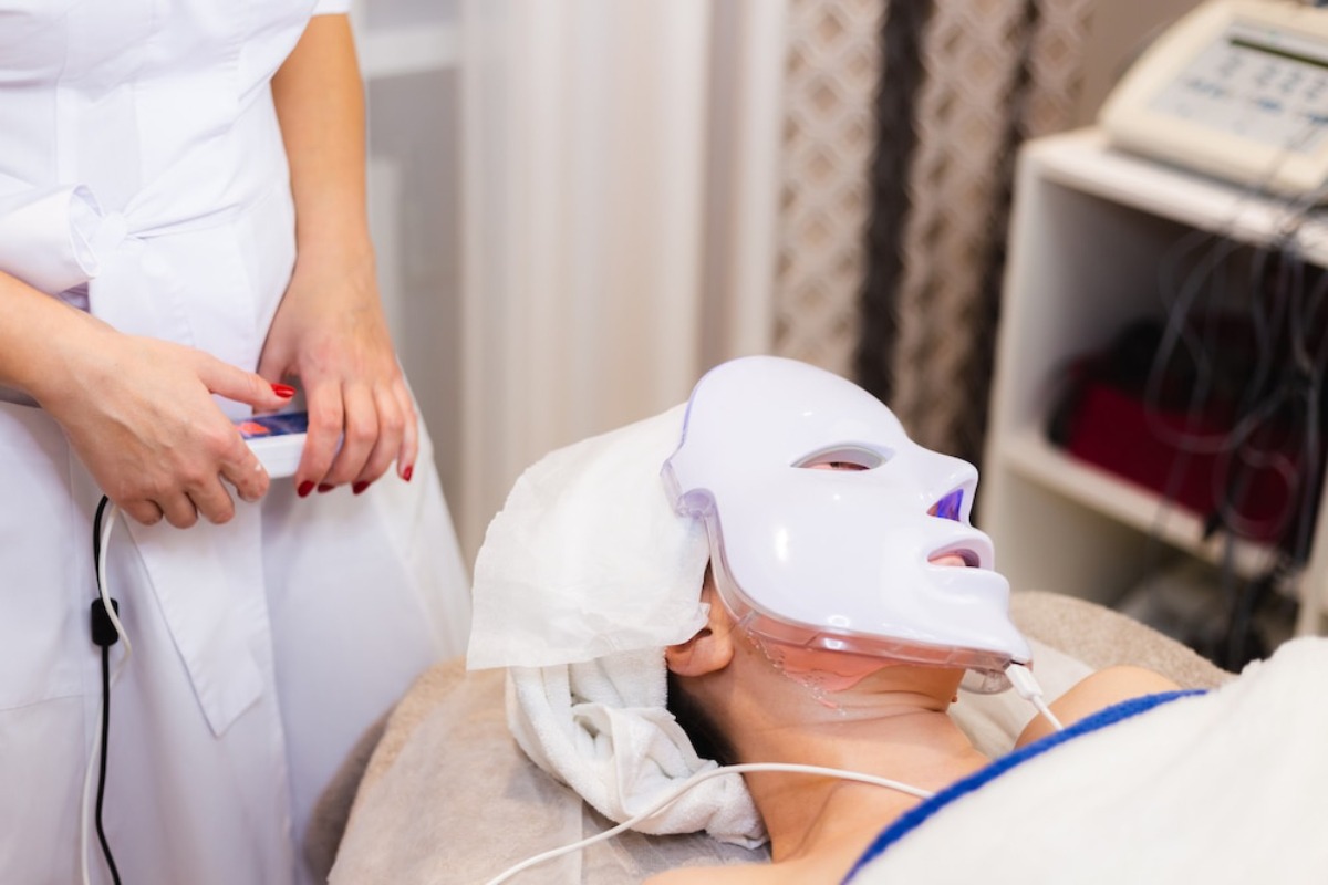A client wearing a LED facial mask receives treatment, while an aesthetician prepares equipment nearby in a serene spa setting.