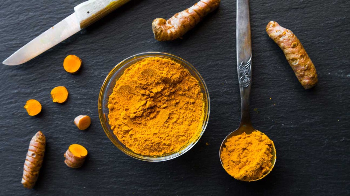 A bowl of bright yellow turmeric powder surrounded by fresh turmeric roots and slices, with a spoonful of powder and a knife nearby.