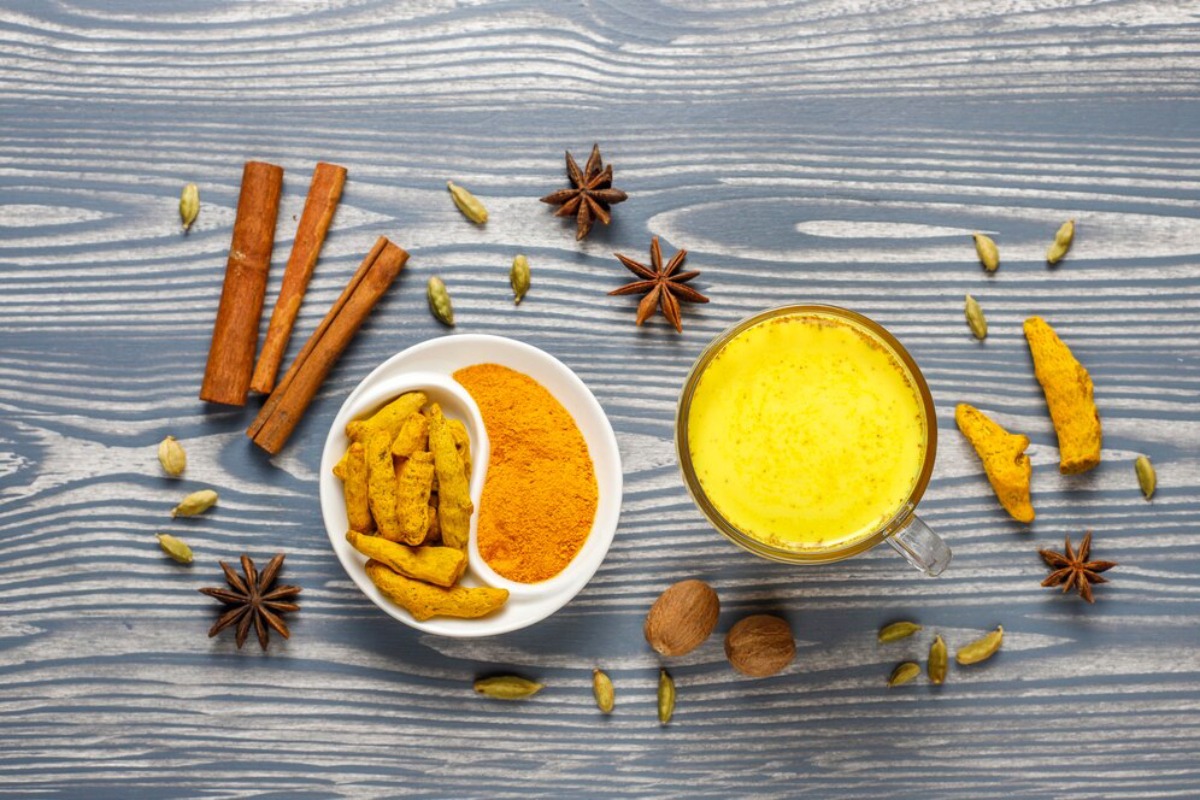 A cup of golden turmeric latte surrounded by various spices: turmeric roots, cinnamon sticks, star anise, and cardamom on a wooden surface.