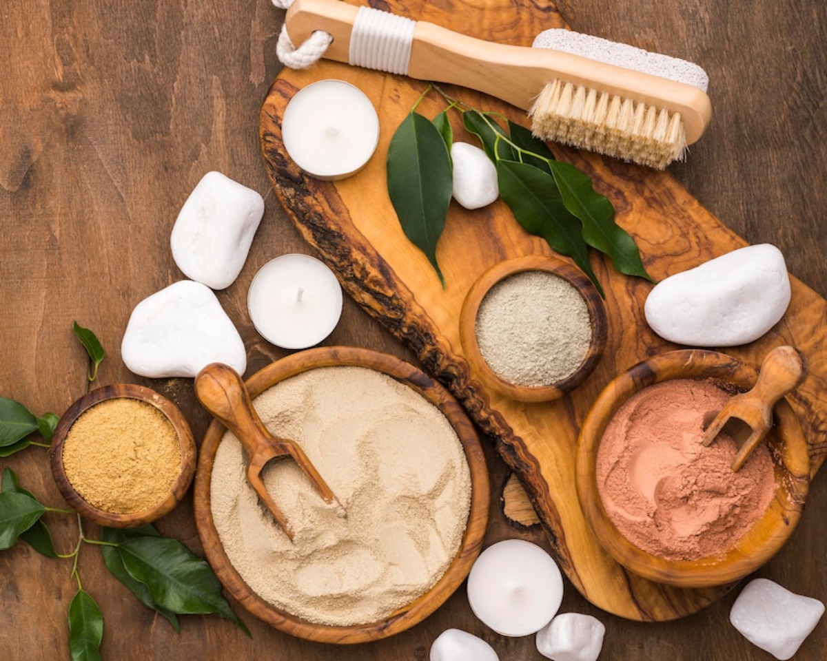 A top-down view of natural powders in wooden bowls, stones, candles, and a brush on a rustic wooden surface, evoking a calming spa vibe.
