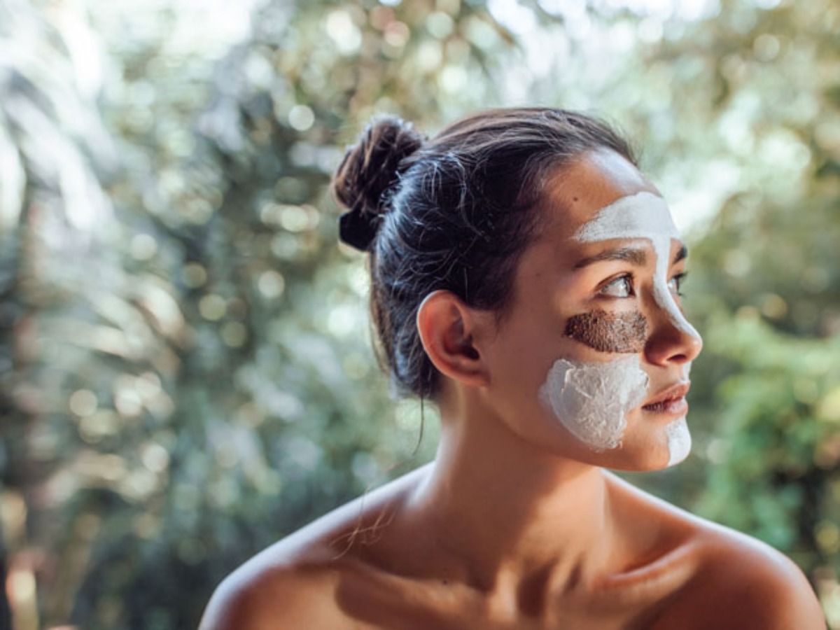 A woman with a bun hairstyle gazes thoughtfully outdoors, surrounded by lush greenery and dappled sunlight.