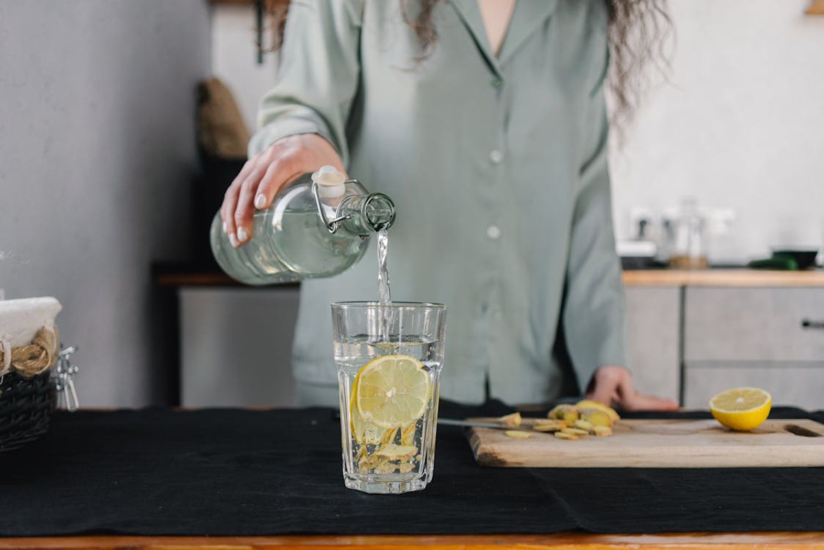 A person in a light green shirt pours water from a glass bottle into a clear glass with lemon slices, on a kitchen countertop.