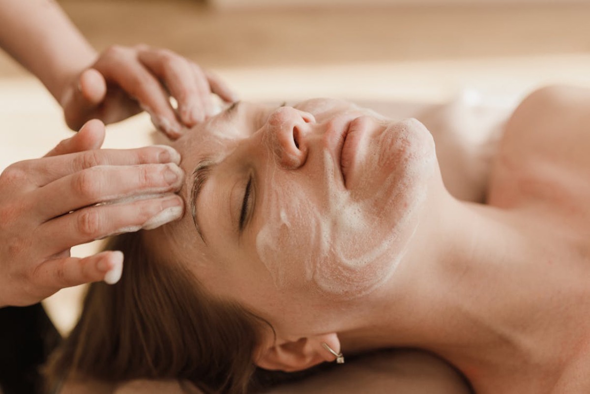 A close-up of hands applying a treatment to the scalp of a person lying down, suggesting a relaxing spa or massage atmosphere.
