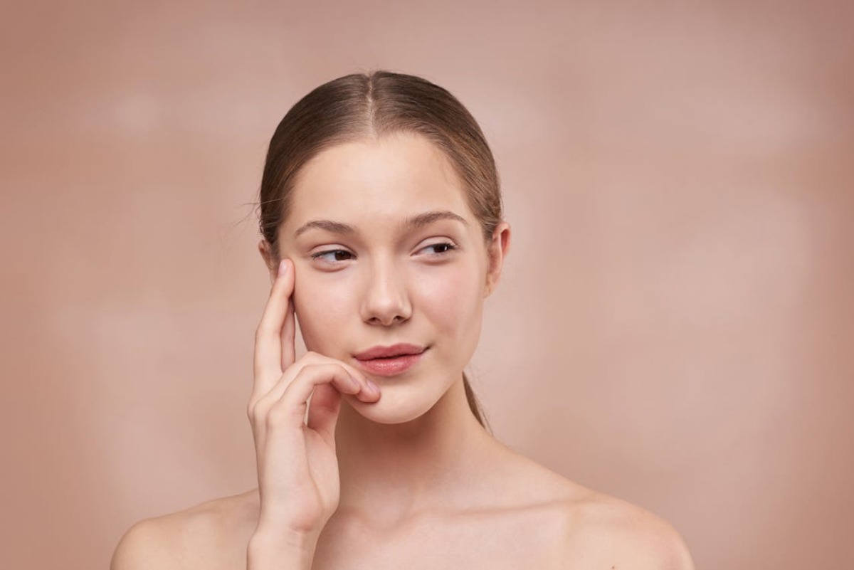 A person with shoulder-length hair poses against a soft, neutral background, thoughtfully touching their cheek.