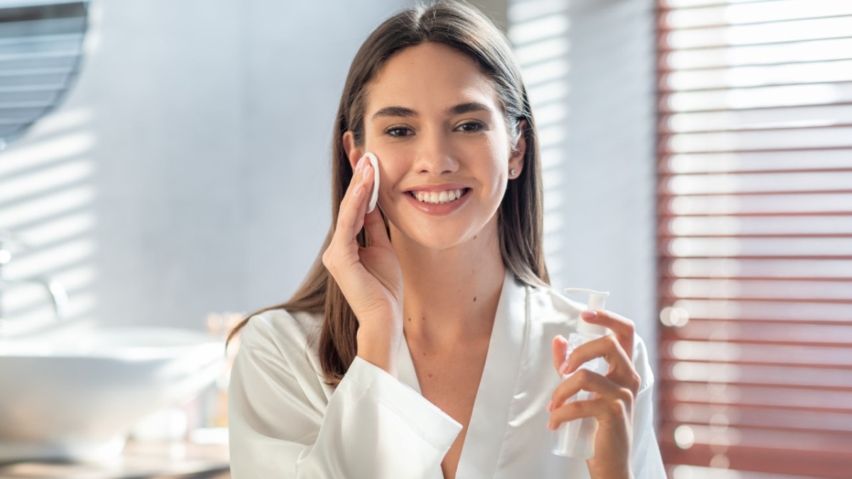 A woman in a white robe holds a skincare product while in a bright, modern bathroom with sunlight streaming through blinds.