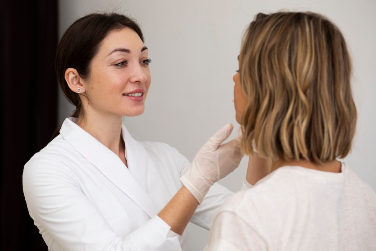 A professional applying skincare or cosmetic treatment to a client in a modern, well-lit setting. The client has shoulder-length hair.