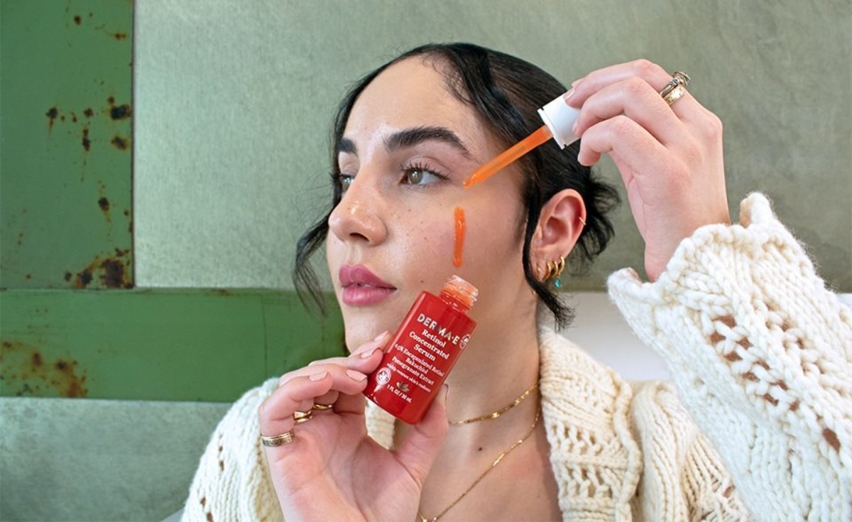 A person applies a dropper of skincare serum while holding a bright red product label, wearing a cozy cream-colored sweater.