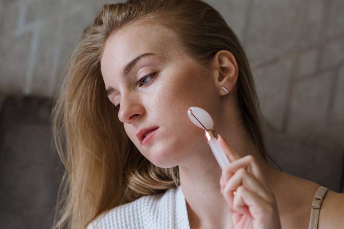 A woman with long blonde hair holds a beauty tool, wearing minimalist silver earrings, dressed in a cozy white sweater.