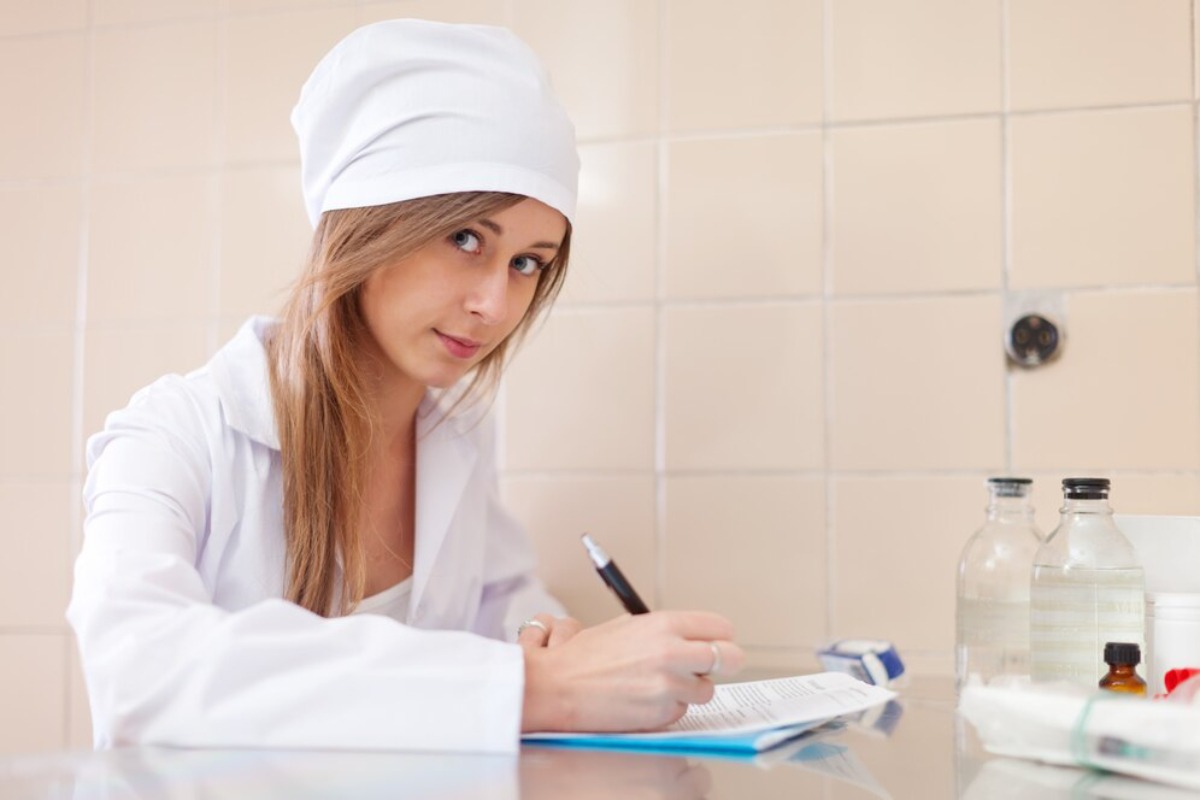 A medical professional in a white coat and hat writes notes at a table with glass vials and medical supplies against a tiled backdrop.