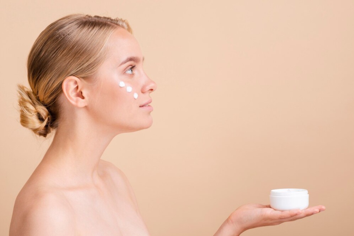A person with blonde hair styled in a bun holds a small white jar against a soft beige background.