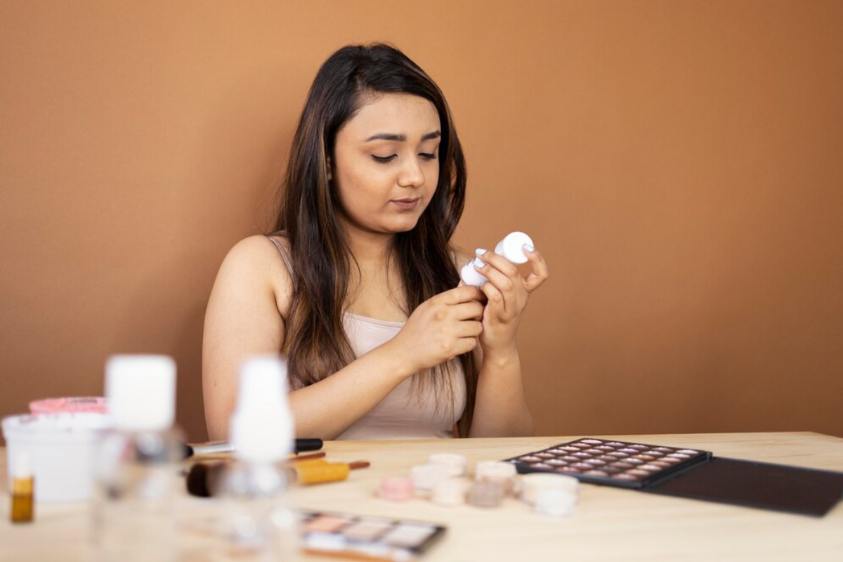A woman with long hair sits at a table, holding a white bottle, surrounded by various makeup products and brushes. Warm brown background.