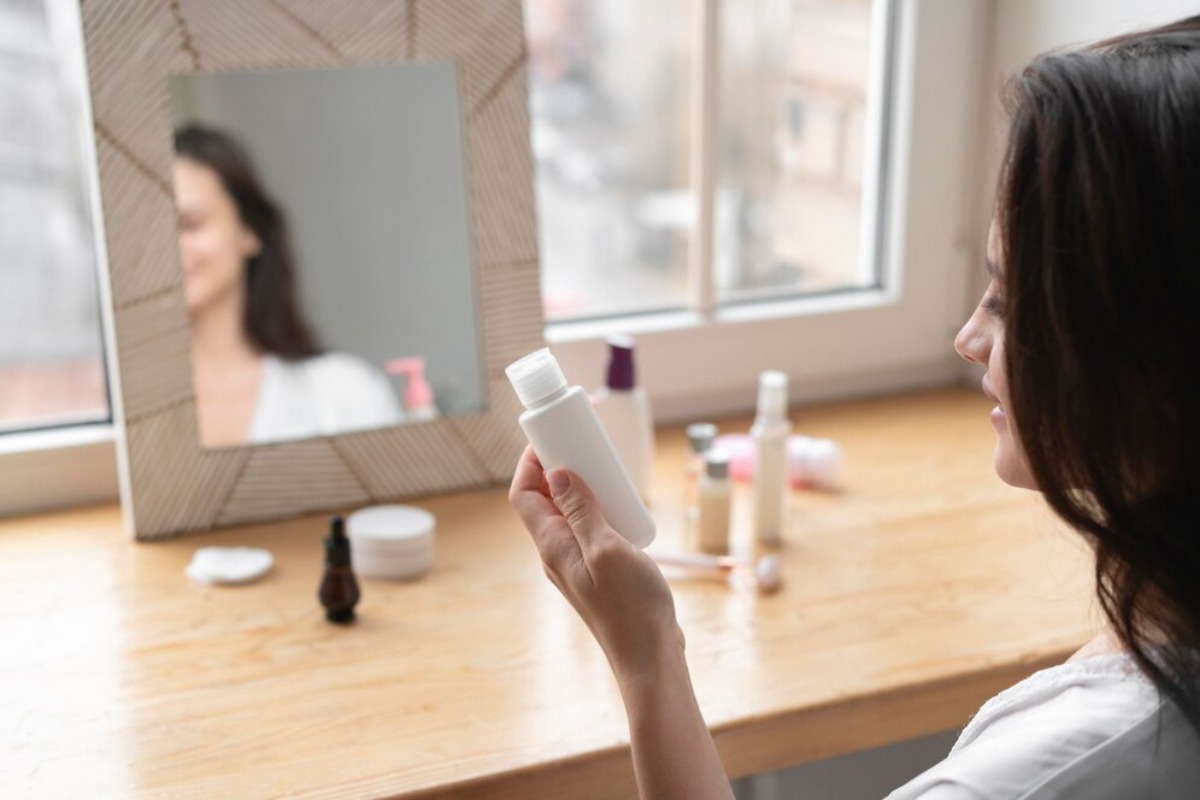 A woman holds a small white bottle while gazing at her reflection in a mirror, surrounded by various skincare products.