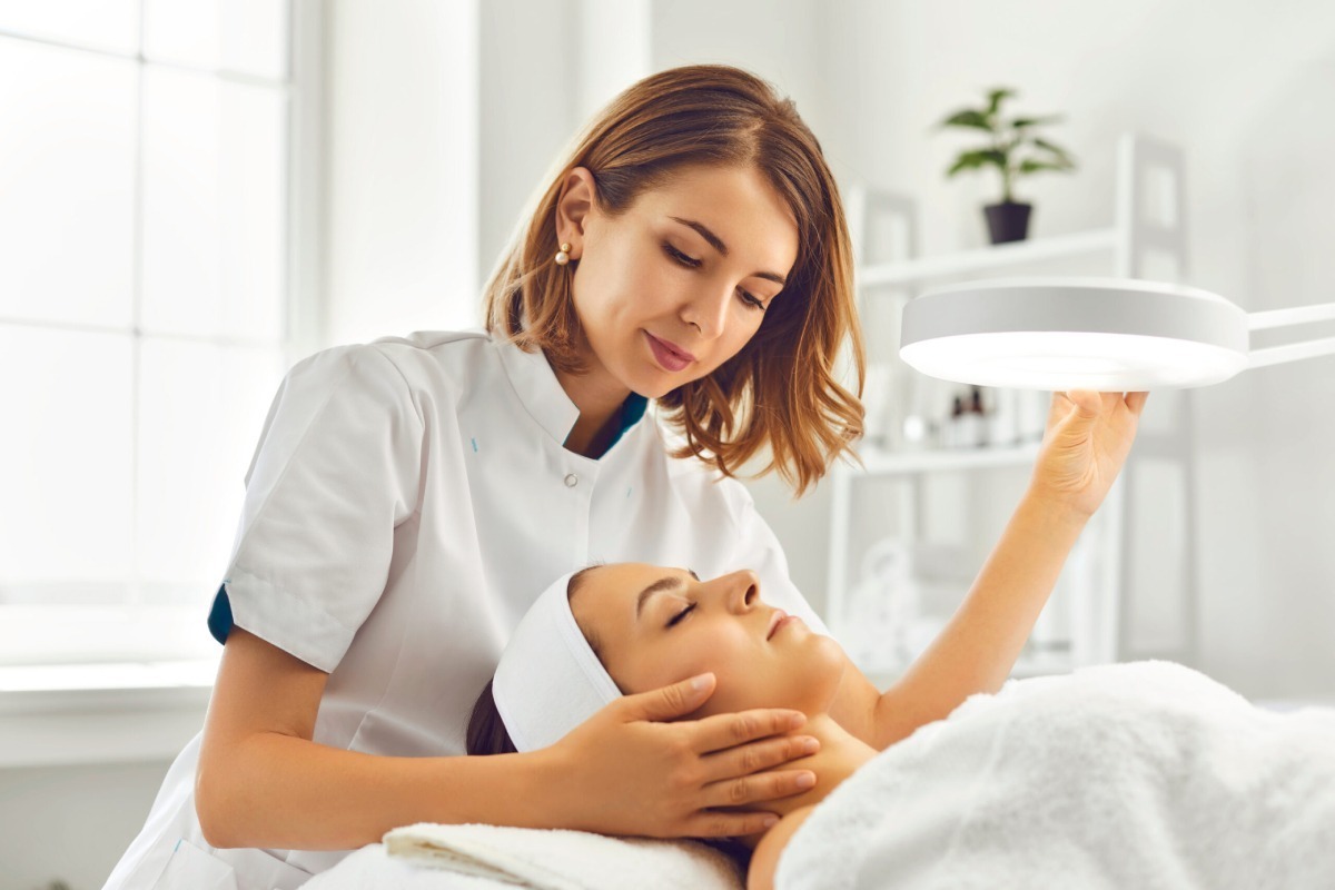 A skincare professional examines a client's facial skin under a bright lamp in a modern treatment room.