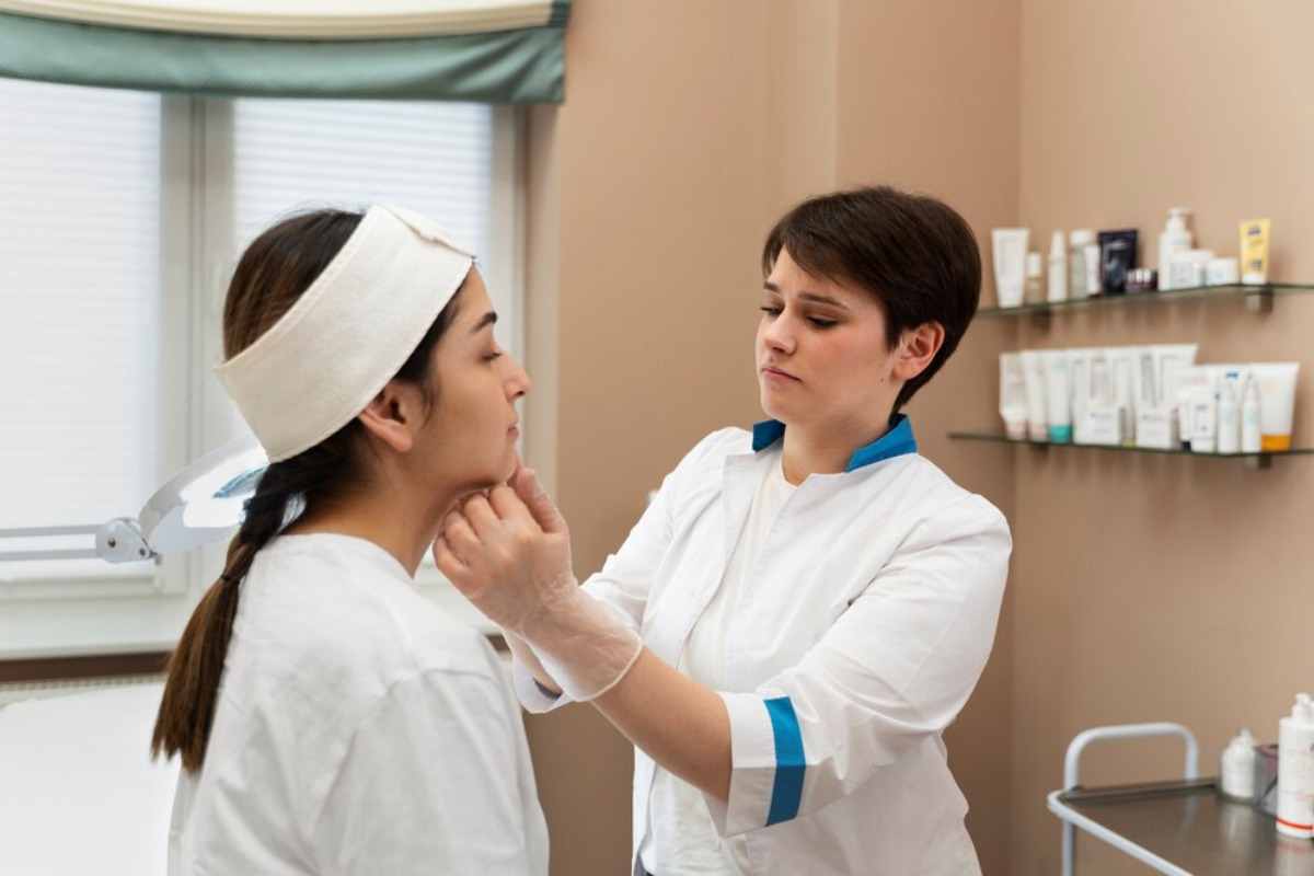 A skincare professional in a white lab coat applies treatment to a client's face in a well-lit clinic, surrounded by skincare products.