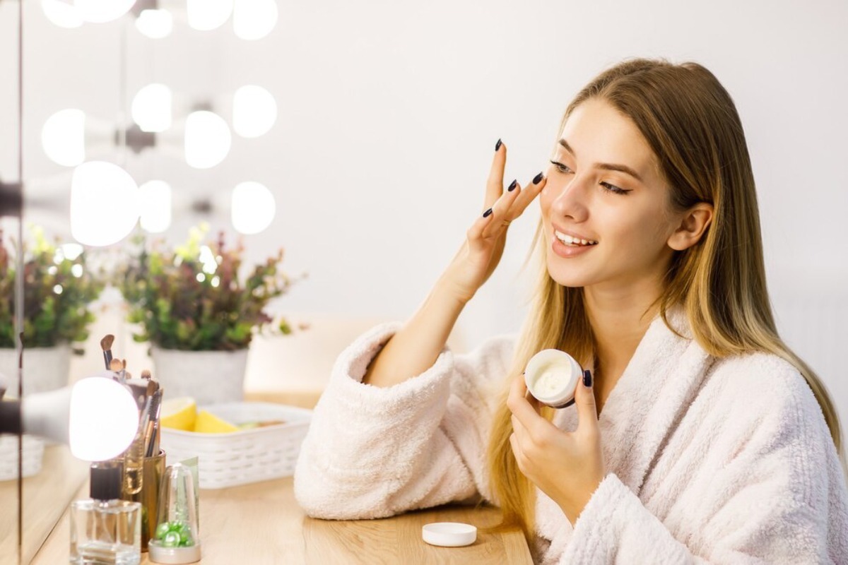 Woman in a white robe applying cream to her face, sitting at a well-lit vanity table.