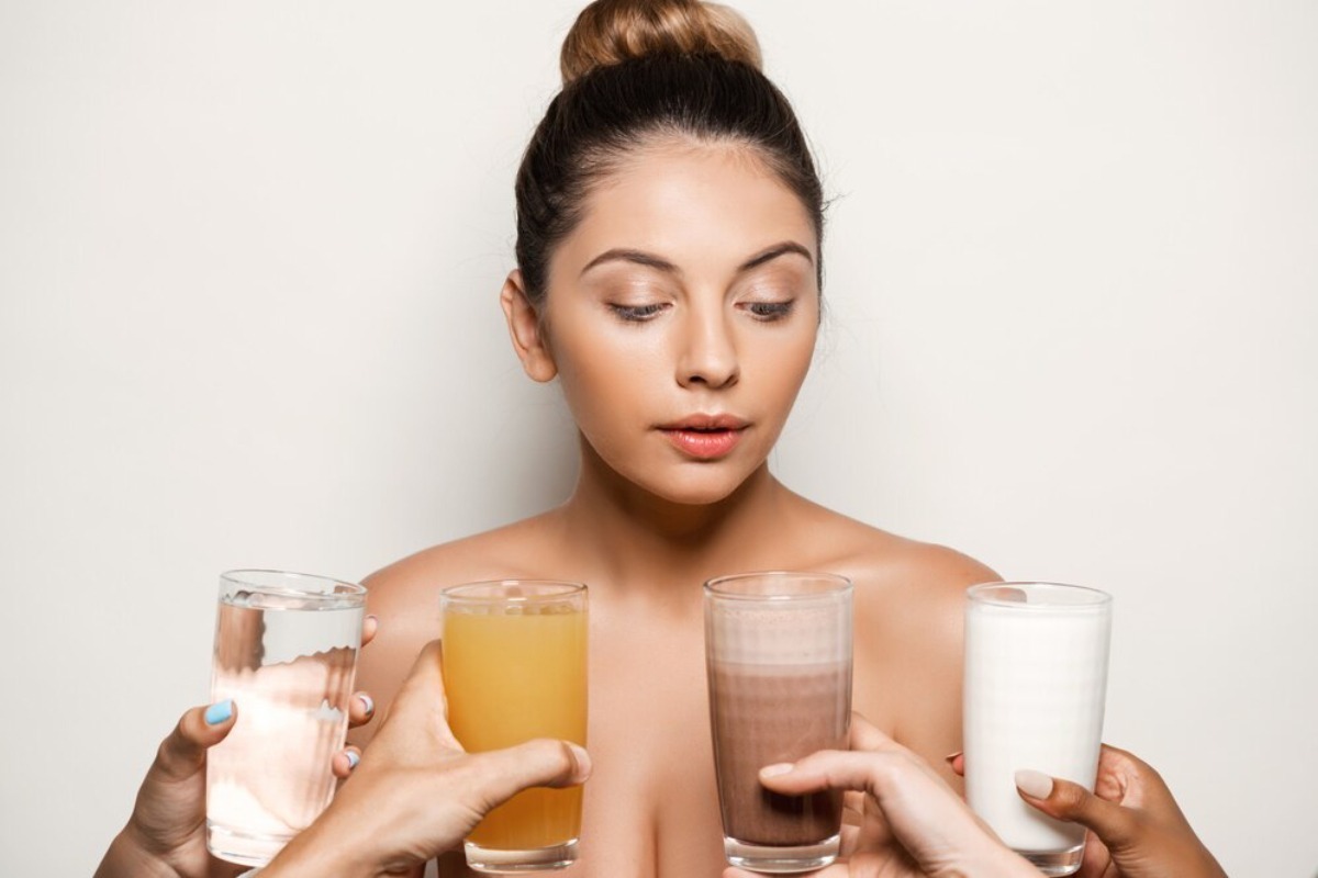 A woman holding four different beverages: clear water, orange juice, chocolate milk, and white milk, against a plain background.