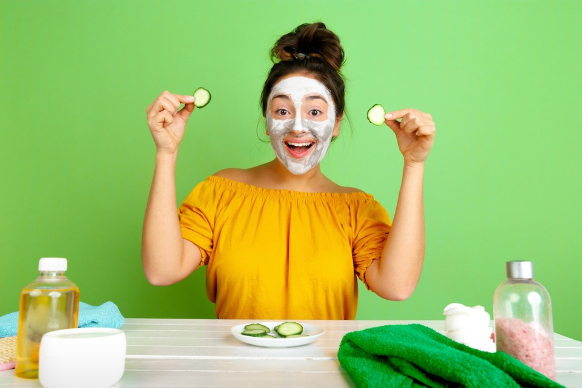 A person wearing a yellow off-shoulder top holds cucumber slices in each hand, surrounded by beauty products on a green background.