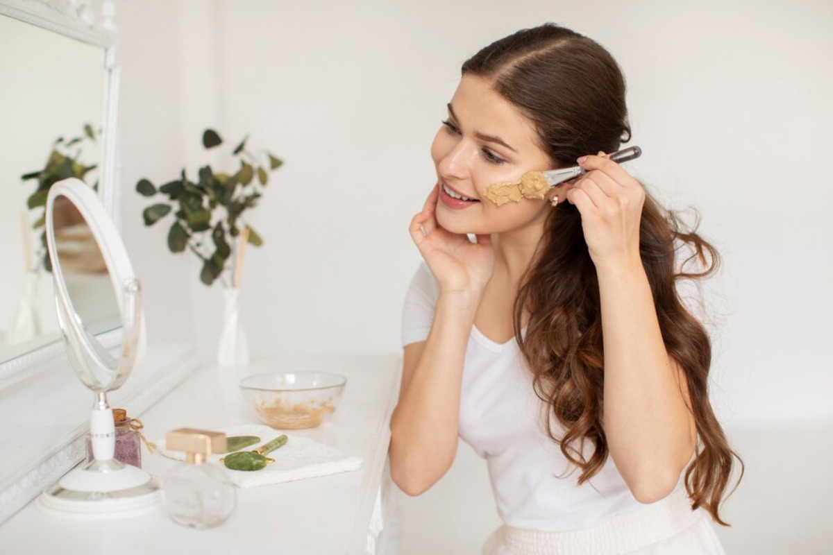 A woman with long, wavy hair is applying makeup at a vanity, surrounded by beauty products and a small potted plant.