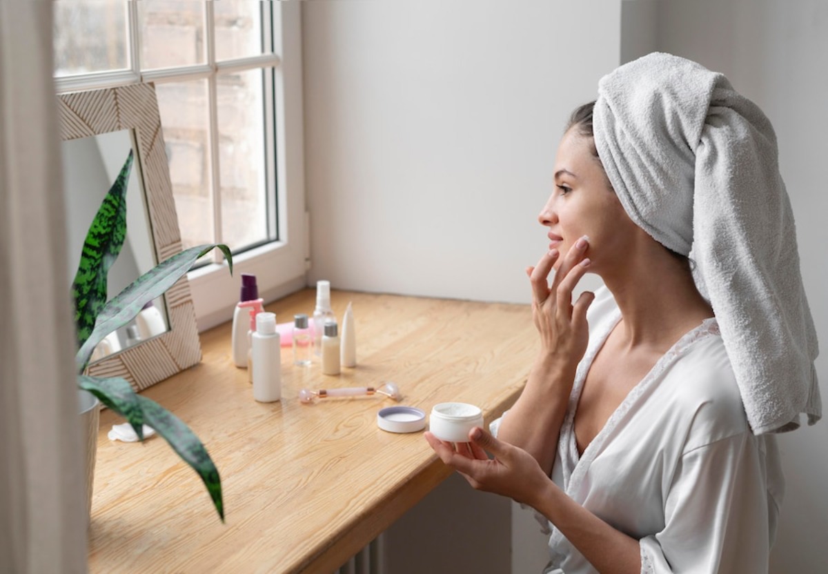 A woman in a robe applies cream at a wooden vanity with various skincare products and a potted plant, illuminated by natural light.