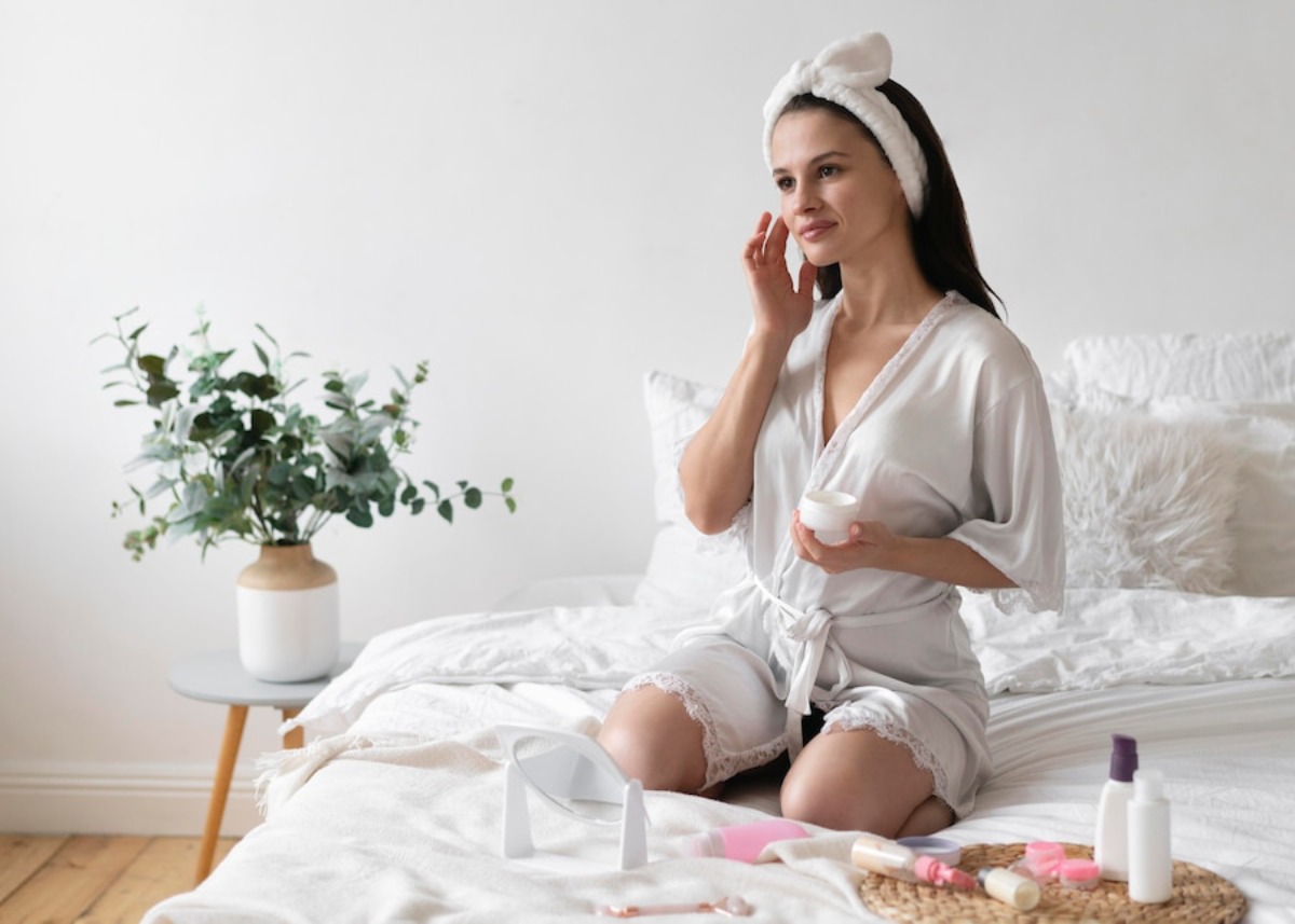 A woman in a light satin robe applies cream while sitting on a bed surrounded by skincare products and a potted plant.
