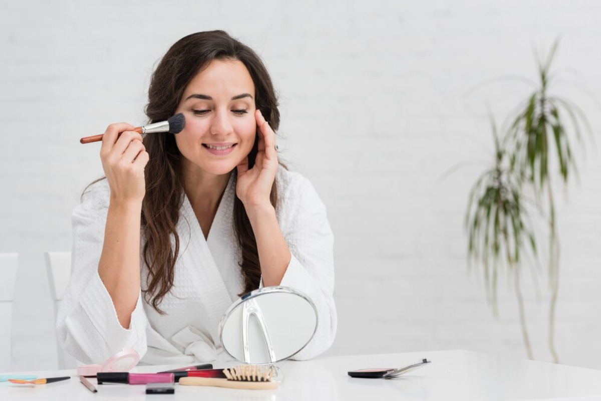 Woman in a white robe applying makeup with a brush, smiling, sitting at a table with cosmetics.