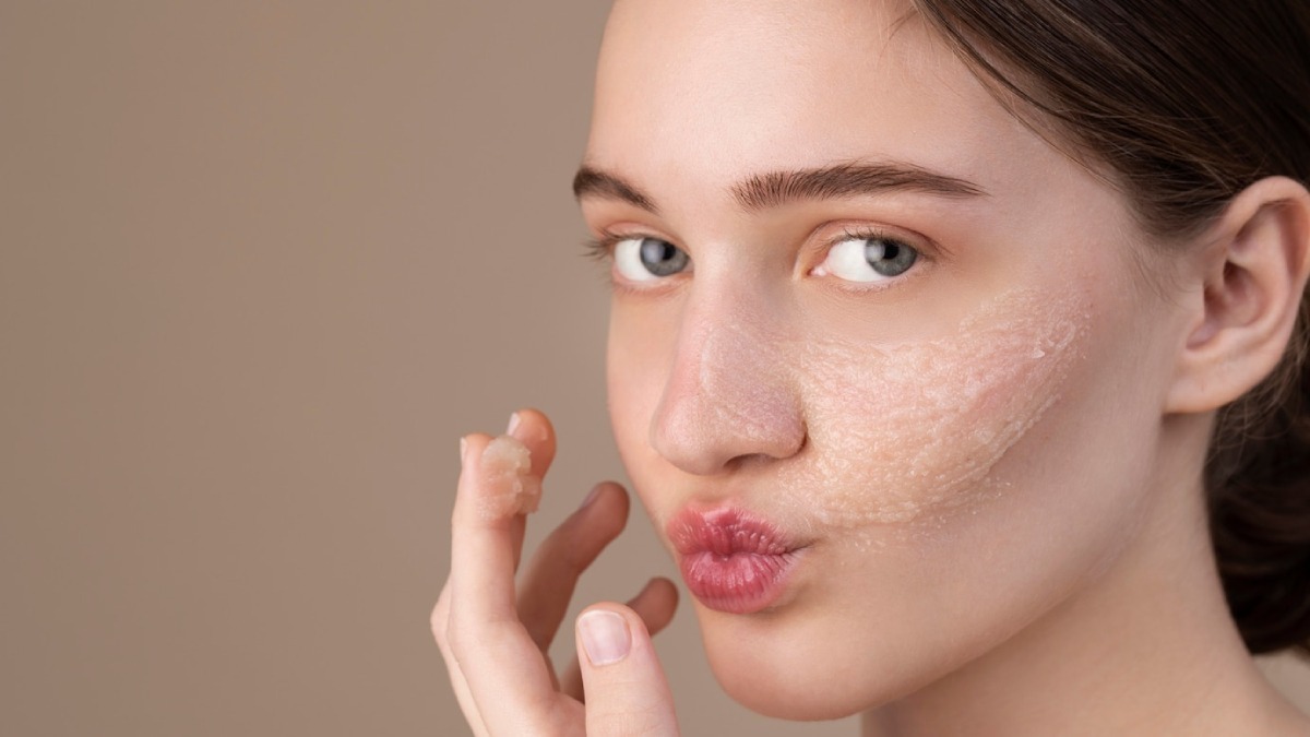 Young woman applying a facial scrub to her cheek, looking at the camera.