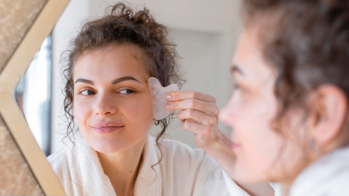 Young woman in a bathrobe using a facial gua sha stone, looking into a mirror.