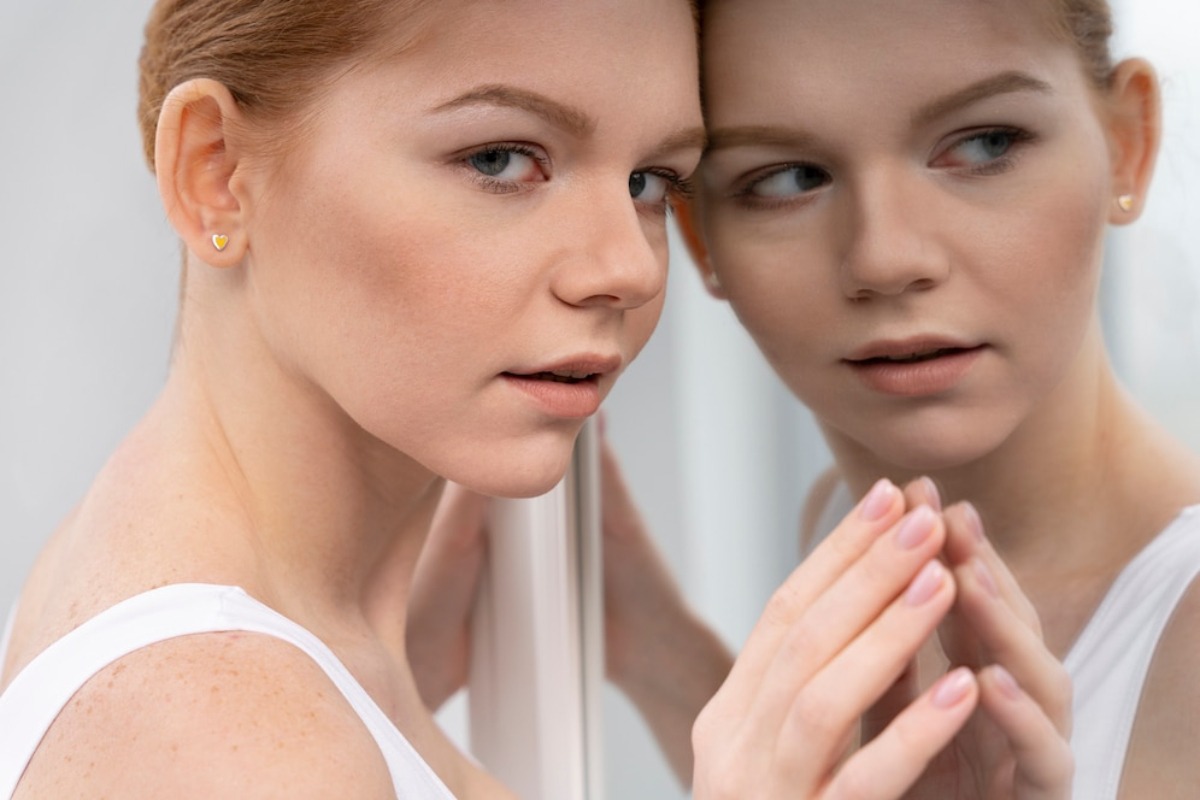 Young woman with red hair looking at her reflection in a mirror, focusing thoughtfully on her face.