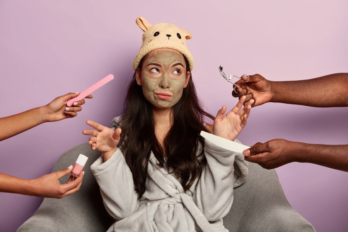 A person in a cozy robe and animal-themed hat is surrounded by hands holding beauty tools and nail polish against a purple backdrop.